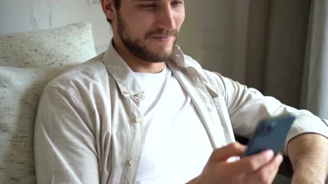 Young Man Uses Smartphone while Sitting on a Sofa, Browses Through Internet Stock Footage 154690798