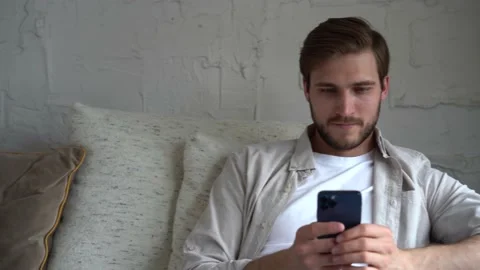 Young Man Uses Smartphone while Sitting on a Sofa, Browses Through Internet Stock Footage 154690806