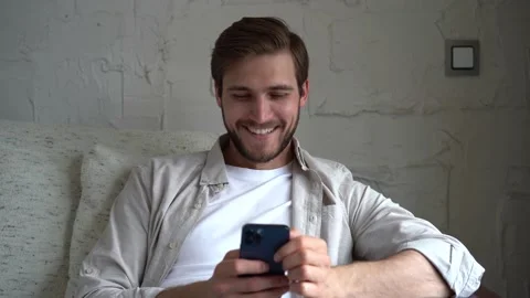 Young Man Uses Smartphone while Sitting on a Sofa, Browses Through Internet Stock Footage 154690816