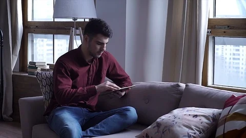 Young man uses tablet with while sitting on sofa at home Stock Footage 105313832