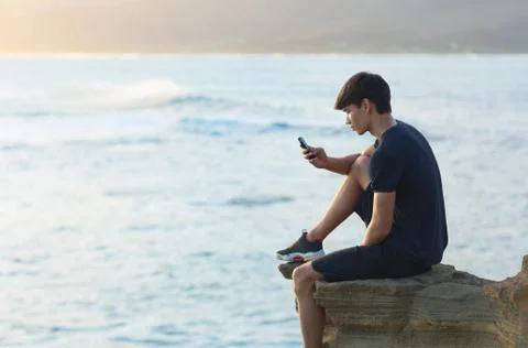 Young man using a cellphone on top a ocean cliff during sunset. Stock Photos