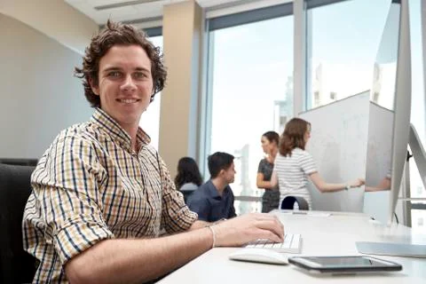 Young man using computer in office looking towards camera, co-workers having 库存照片