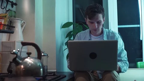 Young man using computer sitting at kitchen while kettle boiling on stove Stock Footage 83871245