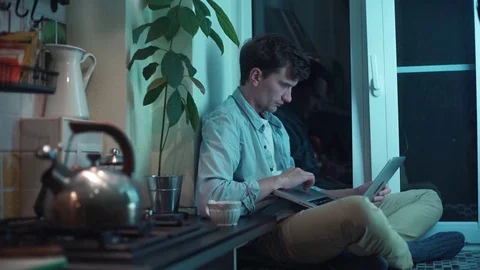 Young man using computer sitting at kitchen while kettle boiling on stove Stock Footage 83871379