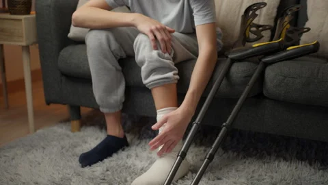 Young man using crutches and massaging his injured leg while sitting on a c.. 스톡 동영상 340026717