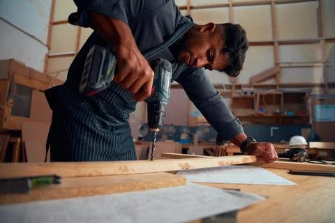 Young man using drill on wood plank during work at woodworking factory Stock Photos