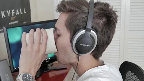 A young man using headphones and drinking coffee on a computer Stock Footage 75487984