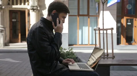 Young man using his laptop on a street bench and talking on the Smartphone Stock Footage 62931812