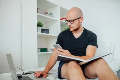 Young man is using his laptop and mobile phone with notepad on his knees. Hom Stock Photos