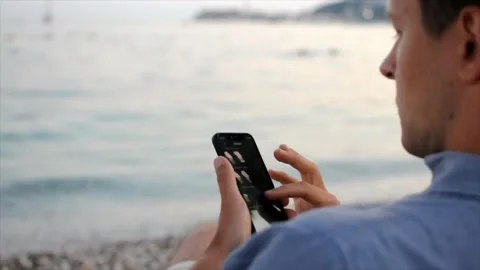 Young man is using his smartphone while sittiing at the beach near the sea Stock Footage 178276365