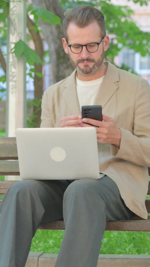 Young Man Using Laptop and Phone while Sitting Outdoor, Vertical Video Stock Footage 319020686