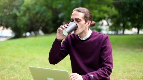 Young man using laptop drinking coffee resting on grass outside in city park. Ur Vidéo 145756238
