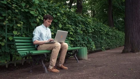 Young man using laptop with headphones sitting on bench at city park. Summer day Stock Footage 77168962