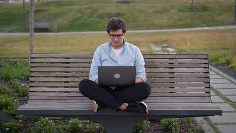 Young man using laptop in park sitting on a bench on blurred grass background 스톡 동영상 118653097