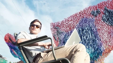 Young man using laptop sitting on chair at city park. Summer day Stock Footage 79356672