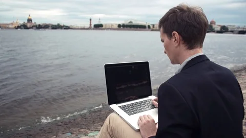Young man using laptop sitting on bench near seafront. Summer day Stock Footage 80063073