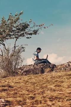 Young man using laptop sitting on a hill against the blue sky Stock Photos