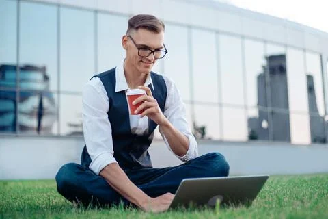 Young man using a laptop sitting on the grass in front of an off Stock Photos