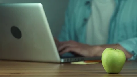 Young man using a laptop typing a message takes a green apple from the table Stock Footage 81882331