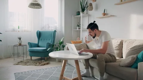 Young man using laptop typing and working sitting on the couch at home and drink Video stock 167285021