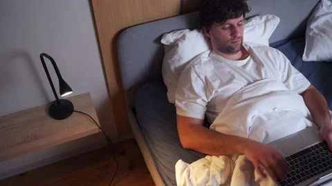 A young man using a laptop while lying on a bed at night. Man is resting in bed 스톡 동영상 233284395
