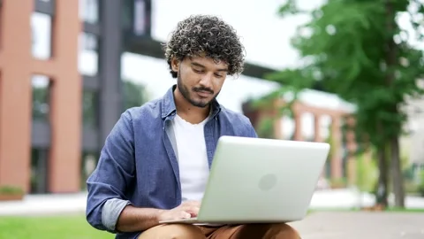 Young man using laptop while sitting on a bench on street near a modern building Stock Footage 302472117