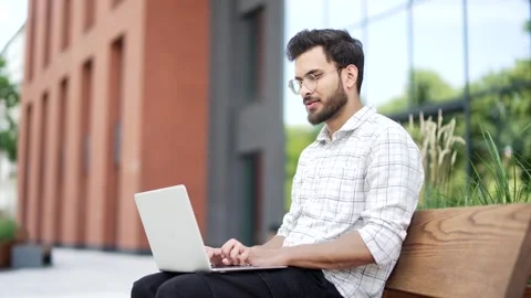 Young man using laptop while sitting on a bench on the street near an modern  Stock Footage 306299560