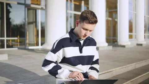Young man using laptop while sitting on steps of modern building during daytime Stock Footage 310752588