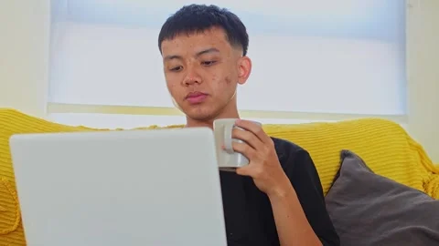 Young man using laptop while drinking warm coffee or tea from mug on yellow sofa Stock Footage 342083847
