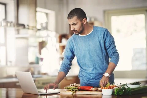 Young man using a laptop while cooking in a kitchen, checking the internet. Male 스톡 사진