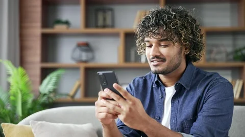 Young man using mobile phone sitting on sofa in living room at home. Smiling  Stock Footage 296764370