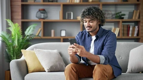 Young man using mobile phone sitting on sofa in living room at home. Smiling  스톡 동영상 334231073