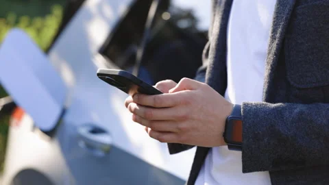 Young man using smartphone near electric car on charging station. Man holding Video stock 249898314