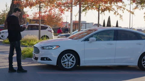 Young man using smartphone near white sedan in urban setting, showcasing modern Stock Footage 317702771