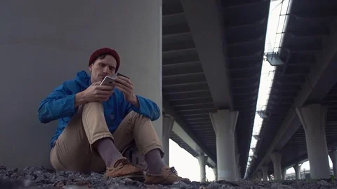 Young man using smartphone sitting on the ground under road bridge. Stock Footage 80285860
