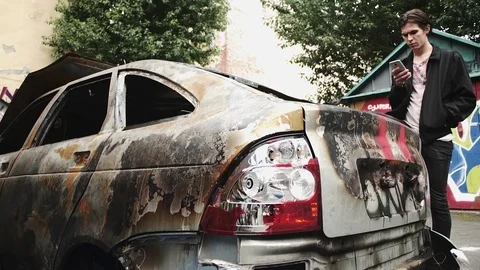 Young man using smartphone standing near crashed burned car. Urban lifestyle Stock Footage 80008527