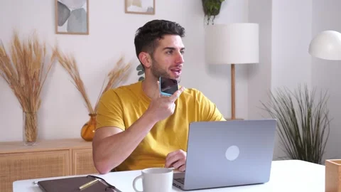 Young man using smartphone while working on laptop at home sending a voice note Stock Footage 231249974