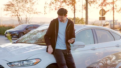Young man using smartphone while leaning against white car in parking lot during Video stock 317703402