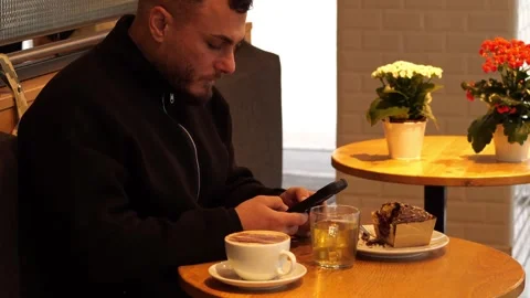 Young Man Using Smartphone While Having Coffee and Cake in Cozy Cafe Vídeos de archivo 332553991