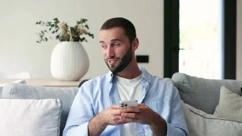 Young man using smartphone while sitting on sofa Vídeos de archivo 335588157