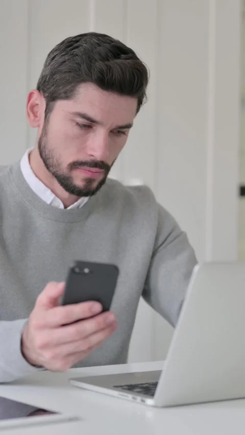 Young Man using Smartphone while using Laptop in Office, Vertical Video Video stock 339582108
