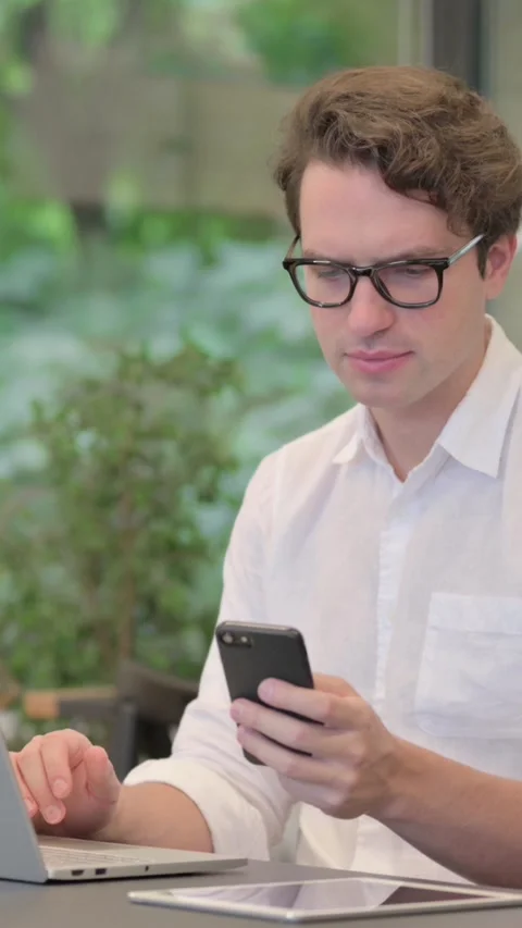 Young Man Using Smartphone while Using Laptop in Modern Office, Vertical Video Stock Footage 340032331