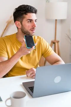 Young man using smartphone while working on laptop at home sending a voice note Stock Photos