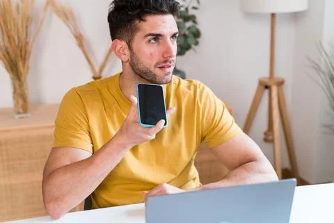 Young man using smartphone while working on laptop at home sending a voice note Stock Photos