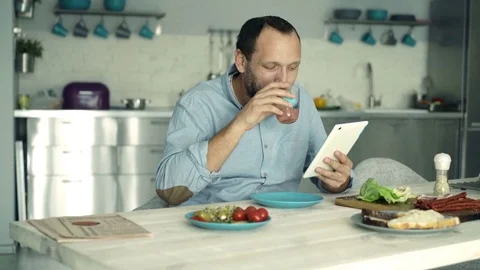 Young man using tablet computer and eating sandwich in kitchen, 4K Stock Footage 74159813