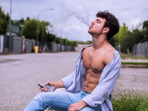 Young man vapes while sitting on pavement in urban area Stock Photos