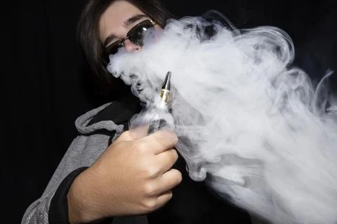 Young man vaping e-cigarette and blowing a cloud on black background. 库存照片