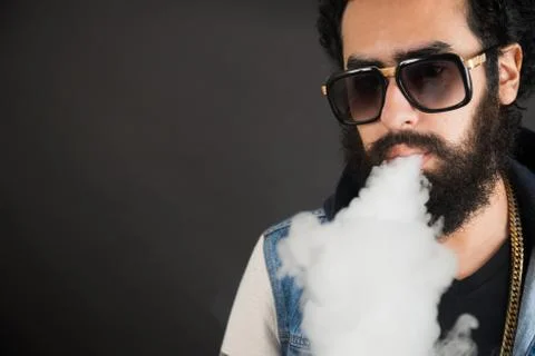 Young man vaping, studio shot. Bearded guy with sunglasses blowing a cloud of Stock Photos
