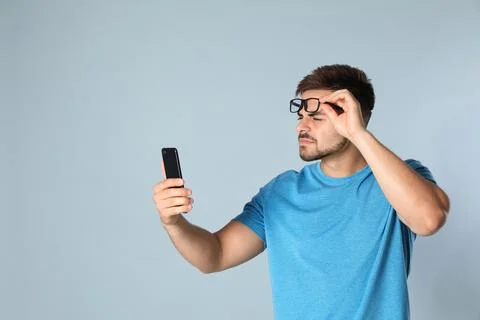 Young man with vision problems using smartphone on grey background, space for Stock Photos