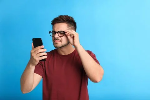 Young man with vision problems using smartphone on blue background, space for Stock Photos